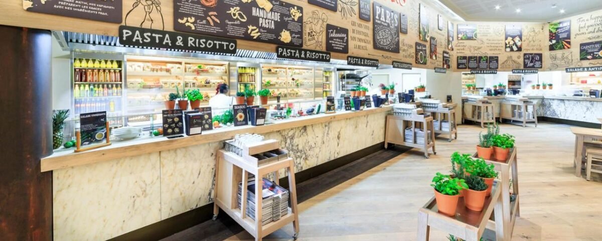 Intérieur d'une cafétéria spacieuse et moderne comprenant un long comptoir avec des panneaux de menu au-dessus, affichant diverses options de restauration comme les pâtes et le risotto de Vapiano. Le décor comprend des plantes en pot et un espace chaleureux et accueillant.