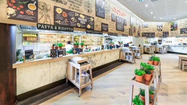Intérieur d'une cafétéria spacieuse et moderne comprenant un long comptoir avec des panneaux de menu au-dessus, affichant diverses options de restauration comme les pâtes et le risotto de Vapiano. Le décor comprend des plantes en pot et un espace chaleureux et accueillant.