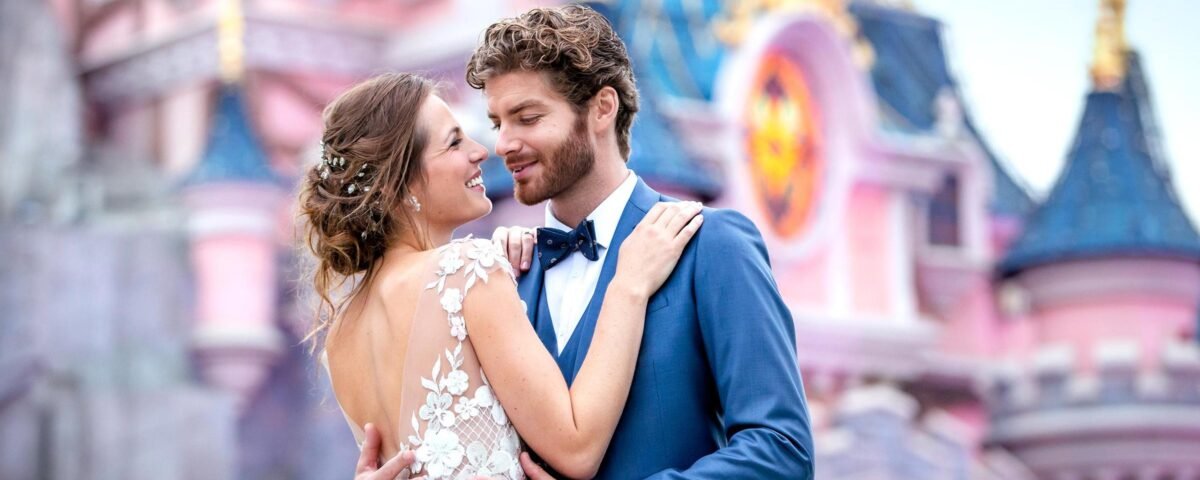 Un couple de jeunes mariés s'enlaçant devant Disneyland Paris, représentant un château de conte de fées. La mariée porte une robe en dentelle, le marié un costume bleu, tous deux souriant joyeusement.