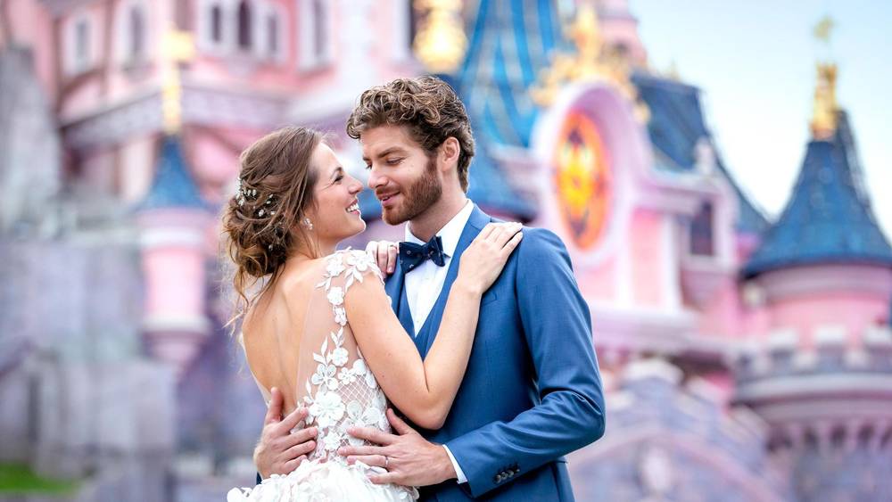 Un couple de jeunes mariés s'enlaçant devant Disneyland Paris, représentant un château de conte de fées. La mariée porte une robe en dentelle, le marié un costume bleu, tous deux souriant joyeusement.