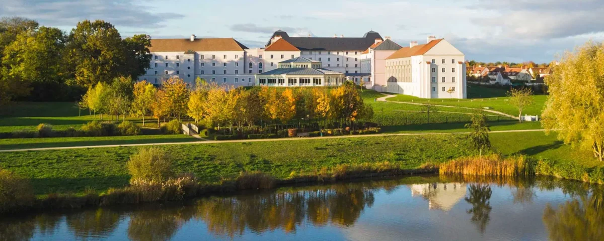 Vue aérienne d'un grand hôtel B&B historique blanc aux toits rouges entouré de pelouses vertes, d'arbres et d'un étang, sous un ciel clair en début de soirée.