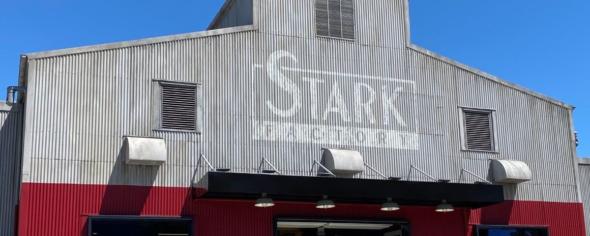 Un bâtiment industriel avec une façade ondulée argentée. "Disneyland Paris" est visiblement embossé sur la partie supérieure, entourée d'un ciel bleu éclatant. Des panneaux rouge foncé encadrent l’entrée en contrebas.