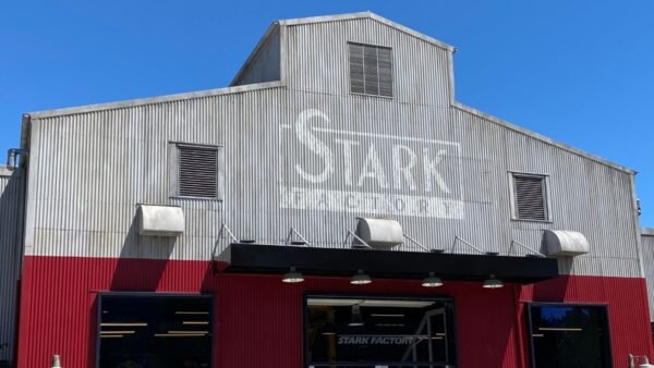 Un bâtiment industriel avec une façade ondulée argentée. "Disneyland Paris" est visiblement embossé sur la partie supérieure, entourée d'un ciel bleu éclatant. Des panneaux rouge foncé encadrent l’entrée en contrebas.