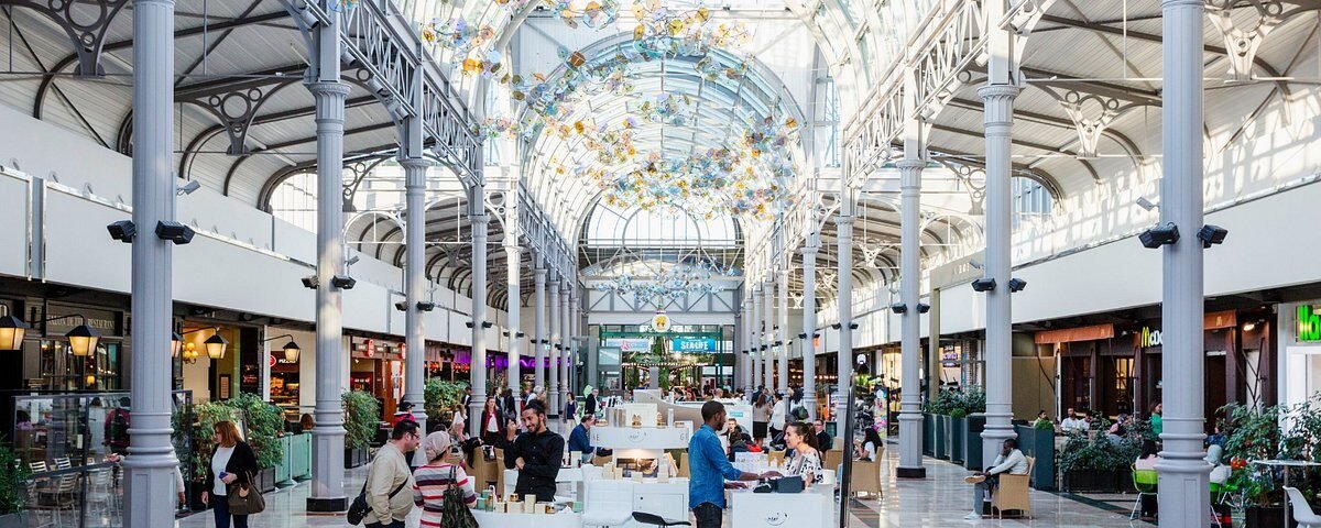 Un centre commercial spacieux et bien éclairé avec un plafond de verre orné, des colonnes métalliques et un café central où les gens se rassemblent et discutent. Des sculptures en verre rappelant Disneyland Paris sont suspendues au-dessus.