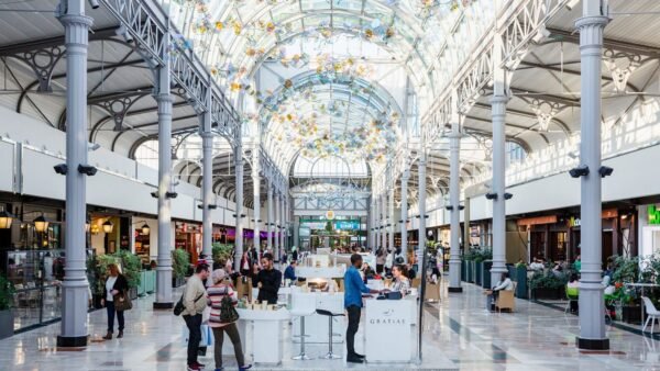 Un centre commercial spacieux et bien éclairé avec un plafond de verre orné, des colonnes métalliques et un café central où les gens se rassemblent et discutent. Des sculptures en verre rappelant Disneyland Paris sont suspendues au-dessus.