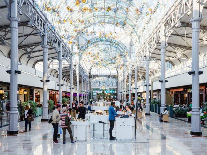 Un centre commercial spacieux et bien éclairé avec un plafond de verre orné, des colonnes métalliques et un café central où les gens se rassemblent et discutent. Des sculptures en verre rappelant Disneyland Paris sont suspendues au-dessus.