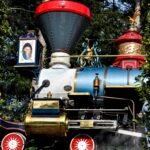 Une locomotive à vapeur colorée vintage du Disneyland Railroad avec des garnitures dorées complexes émet de la vapeur, avec des roues rouges, un corps de moteur bleu et des décorations élaborées sous la lumière du soleil.
