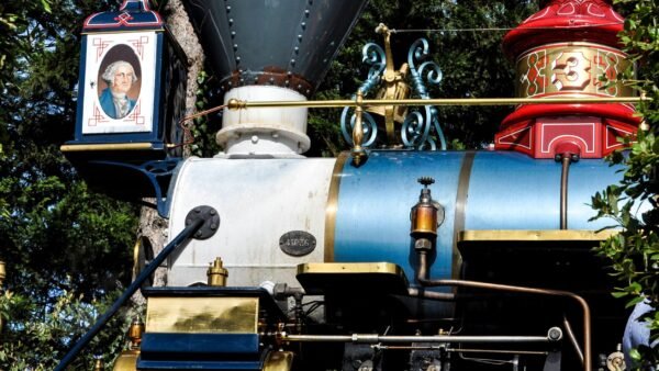 Une locomotive à vapeur colorée vintage du Disneyland Railroad avec des garnitures dorées complexes émet de la vapeur, avec des roues rouges, un corps de moteur bleu et des décorations élaborées sous la lumière du soleil.