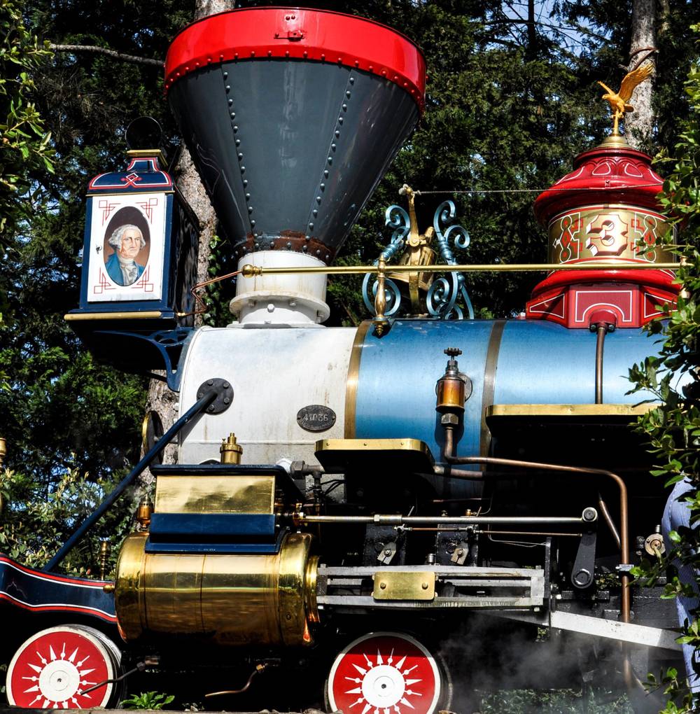 Une locomotive à vapeur colorée vintage du Disneyland Railroad avec des garnitures dorées complexes émet de la vapeur, avec des roues rouges, un corps de moteur bleu et des décorations élaborées sous la lumière du soleil.