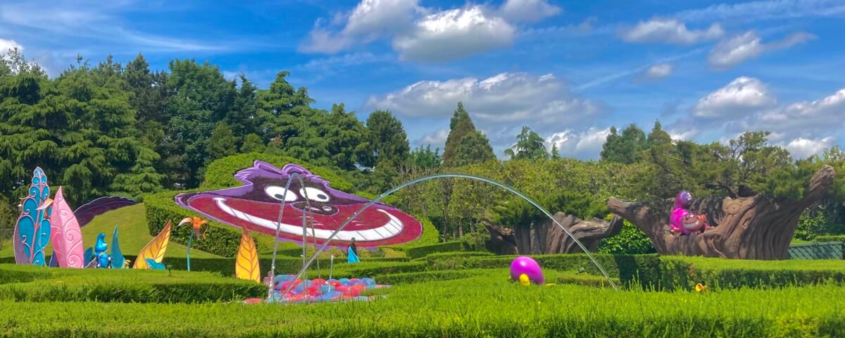 Un jardin coloré sur le thème d'Alice au pays des merveilles avec des sculptures fantaisistes, dont un chat de Cheshire souriant et des fleurs éclatantes, sous un ciel bleu vif avec des nuages duveteux à Disneyland Paris.