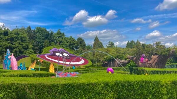 Un jardin coloré sur le thème d'Alice au pays des merveilles avec des sculptures fantaisistes, dont un chat de Cheshire souriant et des fleurs éclatantes, sous un ciel bleu vif avec des nuages duveteux à Disneyland Paris.