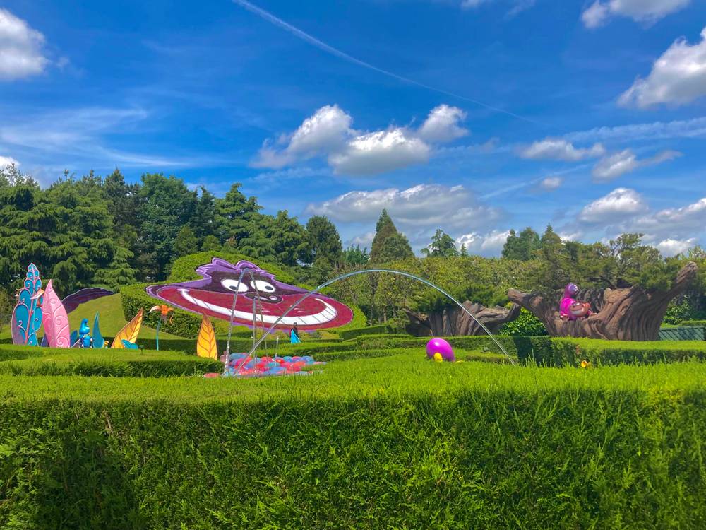 Un jardin coloré sur le thème d'Alice au pays des merveilles avec des sculptures fantaisistes, dont un chat de Cheshire souriant et des fleurs éclatantes, sous un ciel bleu vif avec des nuages duveteux à Disneyland Paris.