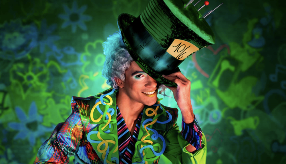 Une personne vêtue d'une tenue fantaisiste, rappelant "Alice au pays des merveilles", brandissant un chapeau surdimensionné avec un fond coloré.