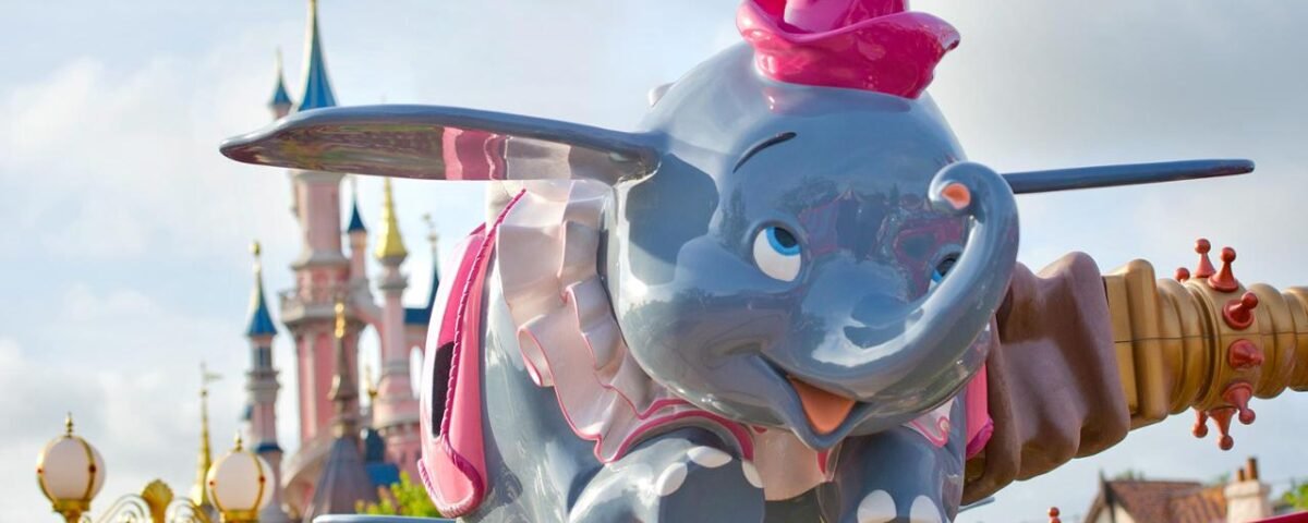 Une statue colorée de Dumbo l'éléphant volant, coiffée d'un chapeau rouge, s'élève sur fond de ciel ensoleillé et des flèches de Disneyland Paris.