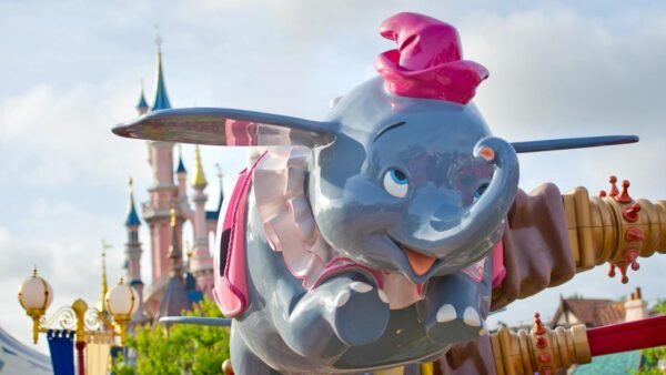 Une statue colorée de Dumbo l'éléphant volant, coiffée d'un chapeau rouge, s'élève sur fond de ciel ensoleillé et des flèches de Disneyland Paris.