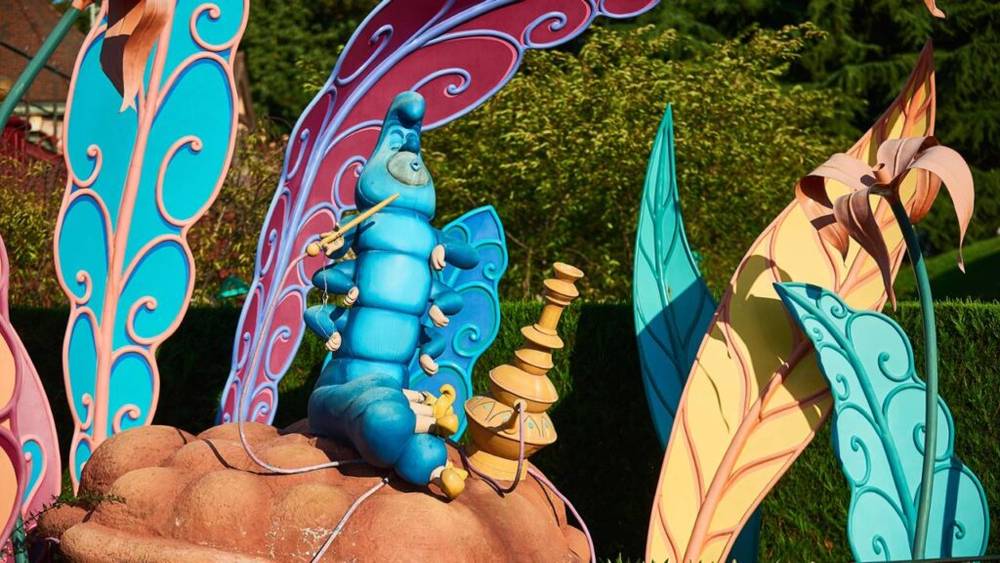 Une statue colorée du personnage chenille d'Alice au pays des merveilles assise sur un champignon, entourée de décorations de jardin fantaisistes à Disneyland Paris.
