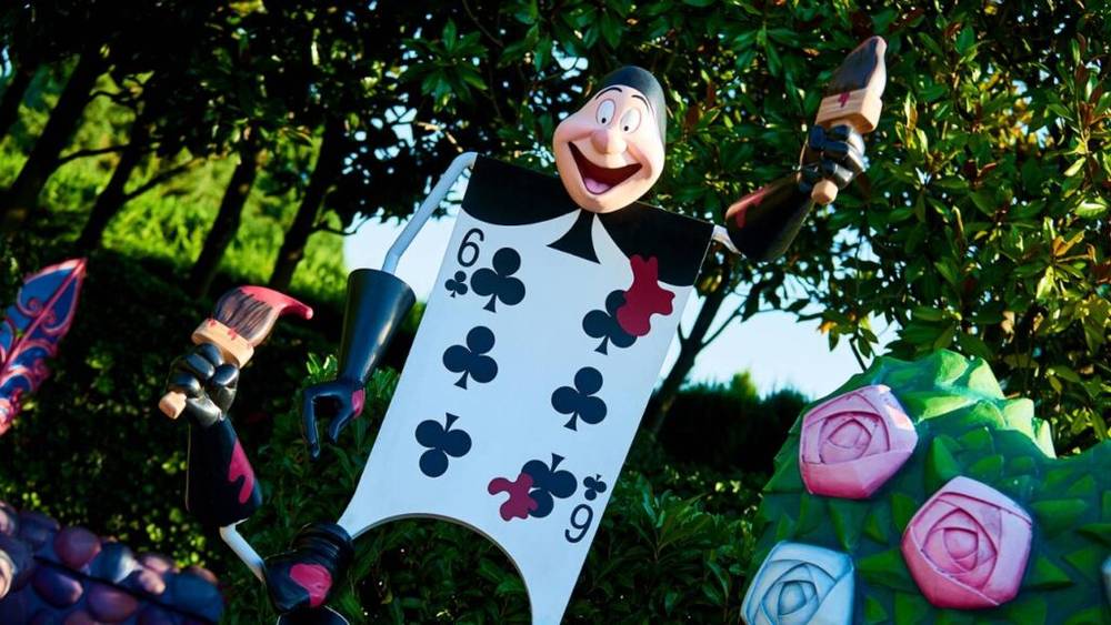 Scène extérieure colorée à Disneyland Paris, avec un personnage de carte à jouer fantaisiste et surdimensionné souriant parmi des roses peintes surdimensionnées et des décorations de jardin du Labyrinthe d'Alice.