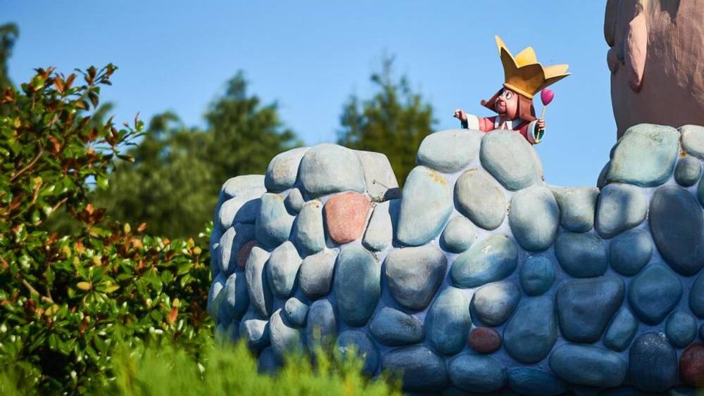 Un personnage haut en couleur en costume de roi au sommet d'un mur de pierre simulé sous un ciel bleu clair, au cœur de l'Etrange Labyrinthe d'Alice à Disneyland Paris.