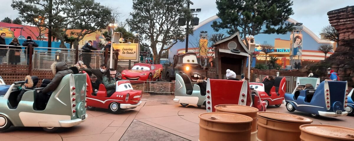 Parc d'attractions sur le thème des voitures à Disneyland Paris, avec des véhicules colorés en forme de voiture avec des visages souriants, parcourant une piste bordée de paysages et de tonneaux caricaturaux, sous un ciel nuageux.