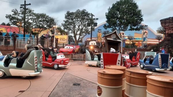 Parc d'attractions sur le thème des voitures à Disneyland Paris, avec des véhicules colorés en forme de voiture avec des visages souriants, parcourant une piste bordée de paysages et de tonneaux caricaturaux, sous un ciel nuageux.