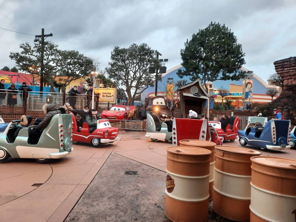 Parc d'attractions sur le thème des voitures à Disneyland Paris, avec des véhicules colorés en forme de voiture avec des visages souriants, parcourant une piste bordée de paysages et de tonneaux caricaturaux, sous un ciel nuageux.