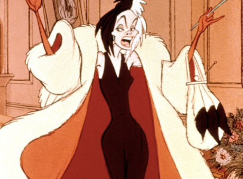 Le personnage animé Cruella de Vil se tient dans une pièce, souriant méchamment, avec ses cheveux noirs et blancs ébouriffés. Elle porte un somptueux manteau de fourrure et une robe rouge, tenant un sac à main et un