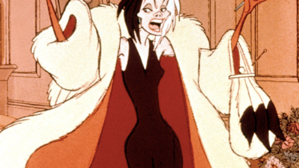 Le personnage animé Cruella de Vil se tient dans une pièce, souriant méchamment, avec ses cheveux noirs et blancs ébouriffés. Elle porte un somptueux manteau de fourrure et une robe rouge, tenant un sac à main et un