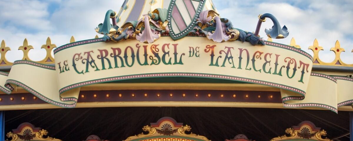 Gros plan sur le dessus d'un carrousel, avec des décorations ornées et l'enseigne "le carrousel de lancelot" en écriture élégante, sur un ciel bleu.
