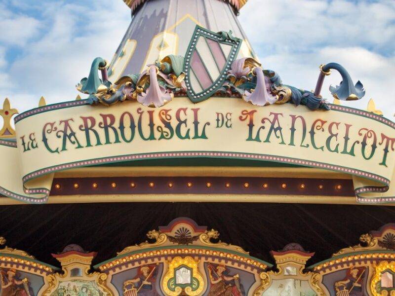 Gros plan sur le dessus d'un carrousel, avec des décorations ornées et l'enseigne "le carrousel de lancelot" en écriture élégante, sur un ciel bleu.