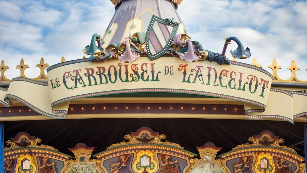 Gros plan sur le dessus d'un carrousel, avec des décorations ornées et l'enseigne "le carrousel de lancelot" en écriture élégante, sur un ciel bleu.