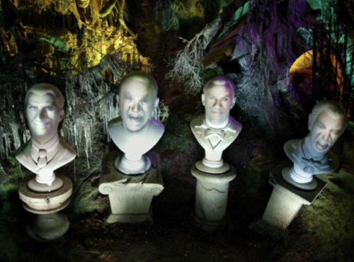 Quatre bustes en pierre avec des visages projetés affichant diverses expressions, sur un fond effrayant et coloré avec des arbres tordus et un éclairage étrange, rappelant les Grim Grinning Ghosts de Phantom Manor.