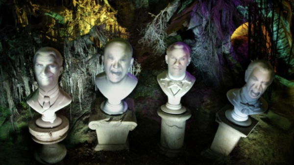 Quatre bustes en pierre avec des visages projetés affichant diverses expressions, sur un fond effrayant et coloré avec des arbres tordus et un éclairage étrange, rappelant les Grim Grinning Ghosts de Phantom Manor.
