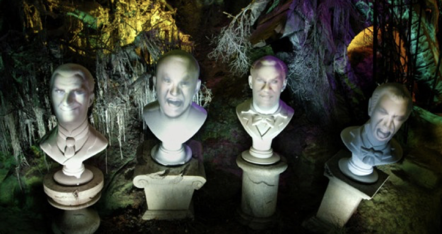 Quatre bustes en pierre avec des visages projetés affichant diverses expressions, sur un fond effrayant et coloré avec des arbres tordus et un éclairage étrange, rappelant les Grim Grinning Ghosts de Phantom Manor.