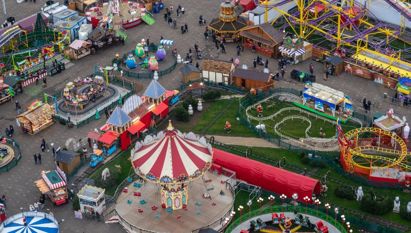 Vue aérienne d'un parc d'attractions coloré avec divers manèges, jeux de carnaval et de nombreuses personnes se promenant, rappelant les meilleurs parcs d'attractions de France.
