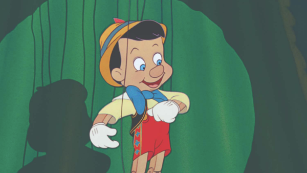 Une marionnette en bois animée ressemblant à Pinocchio de Disney, avec un chapeau jaune, un nœud papillon bleu, un short rouge et des gants blancs, est suspendue à des ficelles sur un fond vert.