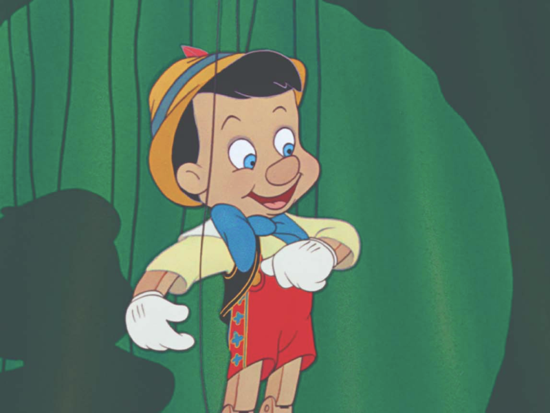 Une marionnette en bois animée ressemblant à Pinocchio de Disney, avec un chapeau jaune, un nœud papillon bleu, un short rouge et des gants blancs, est suspendue à des ficelles sur un fond vert.