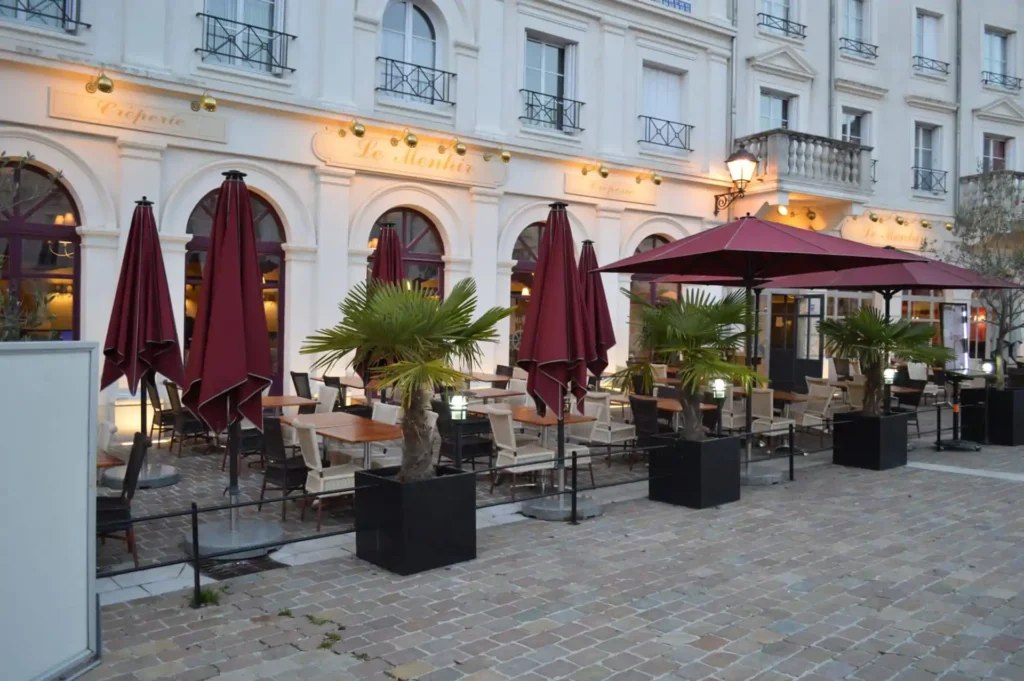 L'espace extérieur du restaurant Goma comprend des parasols rouges, des palmiers et des tables et chaises vides, situés à côté d'un bâtiment avec des fenêtres cintrées et une signalisation française.