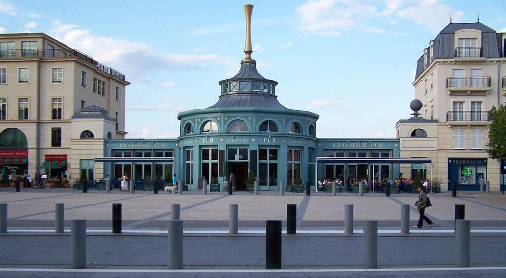 Un bâtiment bleu et blanc avec un dôme unique et une haute structure en forme de corne sur le toit, étiqueté « Brasserie Pub », est situé sur une large place entourée de bâtiments modernes à plusieurs étages à Val d'Europe.