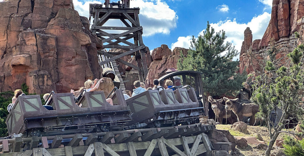 Un groupe de personnes monte sur les montagnes russes du train minier Big Thunder Mountain à travers un décor de parc d'attractions sur le thème du désert rocheux, avec quelques arbres et un ciel bleu en arrière-plan.