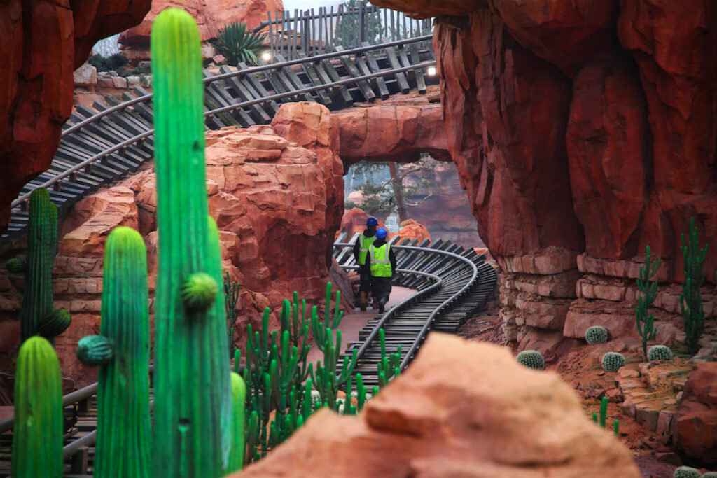 Deux ouvriers en équipement de sécurité marchent le long de la piste des montagnes russes de Big Thunder Mountain, entourés de formations rocheuses rouges et de cactus dans un décor de parc d'attractions.