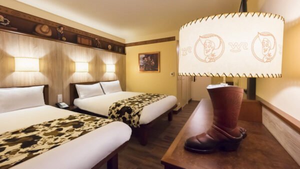 Chambre d'hôtel avec deux lits doubles, plaids à imprimé vache, lampe en forme de botte et abat-jour à thème cow-boy. Des éléments de décoration Disney/Pixar ornent le mur de cet espace charmant inspiré de Cheyenne.