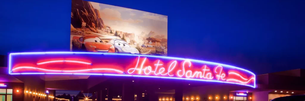 Un panneau au néon indiquant « Disney Hotel Santa Fe » brille sous un grand panneau d'affichage mettant en vedette les personnages d'une animation sur le thème de la voiture la nuit, capturant l'essence de la Route 66 le long du Rio Grande.