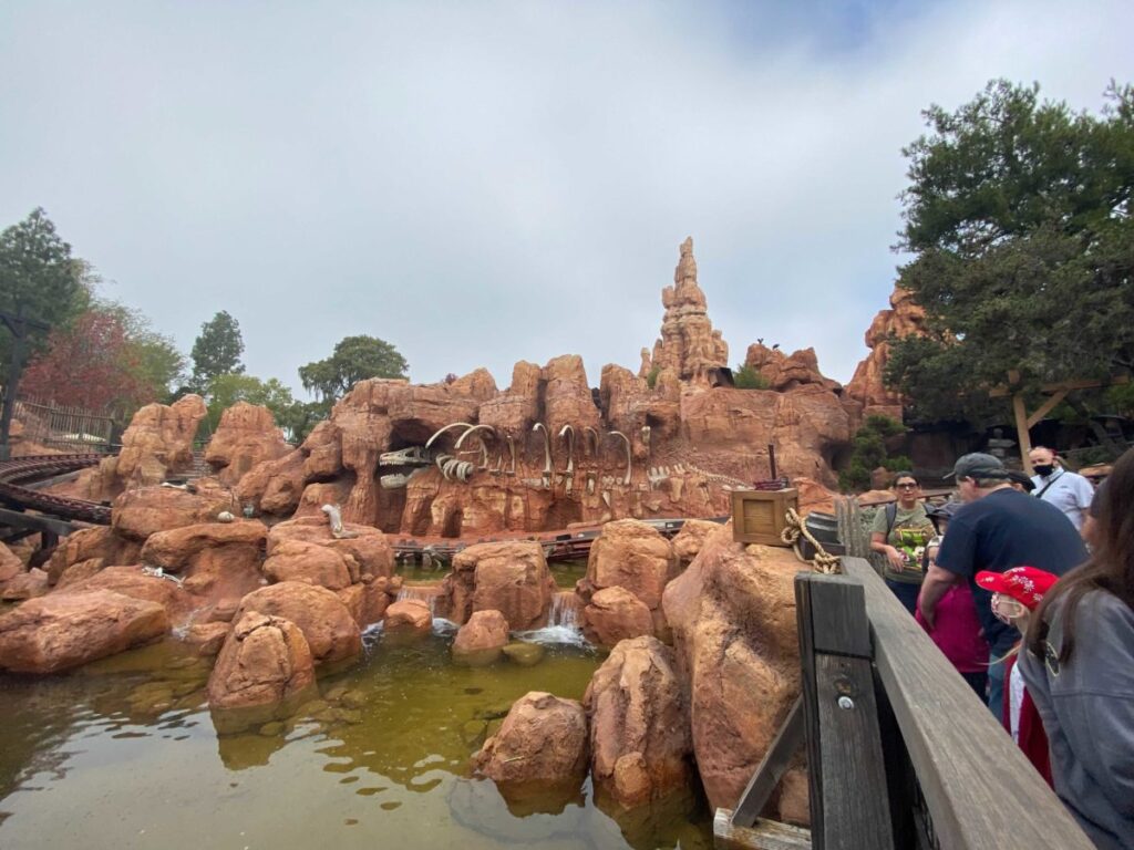 Les gens font la queue près de la structure rocheuse de Big Thunder Mountain, qui présente un squelette de dinosaure, dans un parc d'attractions par temps nuageux.