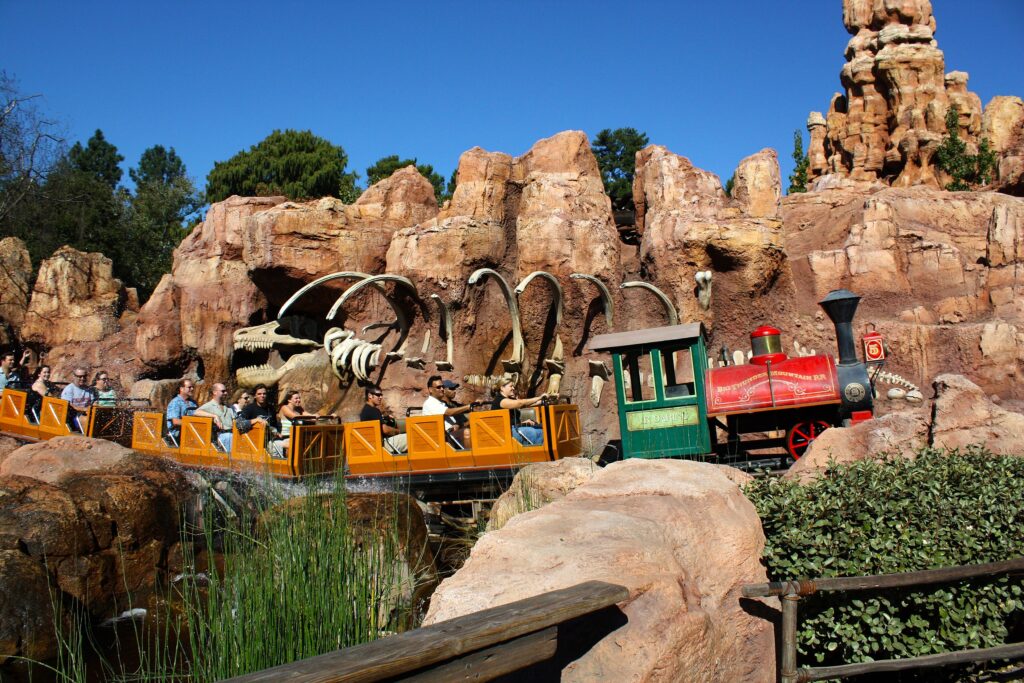 Un train rouge et vert vif sur Big Thunder Mountain traverse un terrain rocheux avec des structures de dinosaures squelettiques, transportant des passagers sous un ciel bleu clair.