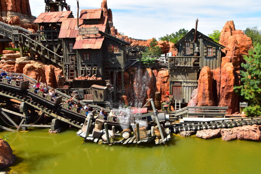 Des montagnes russes dévalent une pente à toute vitesse, éclaboussant l'eau dans un décor de parc d'attractions sur le thème du Far West avec des bâtiments en bois et des structures en roche rouge rappelant Big Thunder Mountain.