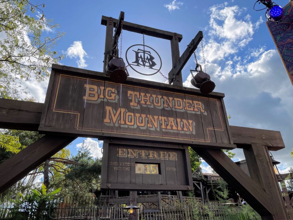 Un panneau en bois indiquant « Big Thunder Mountain » marque l'entrée de l'attraction emblématique, avec un affichage du temps d'attente indiquant 15 minutes, sur fond d'arbres et d'un ciel partiellement nuageux.