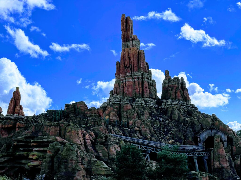 Une montagne rocheuse accidentée, connue sous le nom de Big Thunder Mountain, présente une voie ferrée qui tourne en spirale autour d'elle sous un ciel bleu avec des nuages épars.