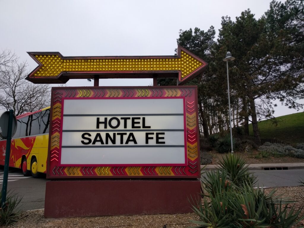 Un panneau avec une flèche jaune et des lumières indique le Disney Hotel Santa Fe, niché le long du Rio Grande. Un bus rouge et jaune est partiellement visible en arrière-plan, ajoutant à la scène animée. Le panneau est entouré d'arbustes luxuriants.