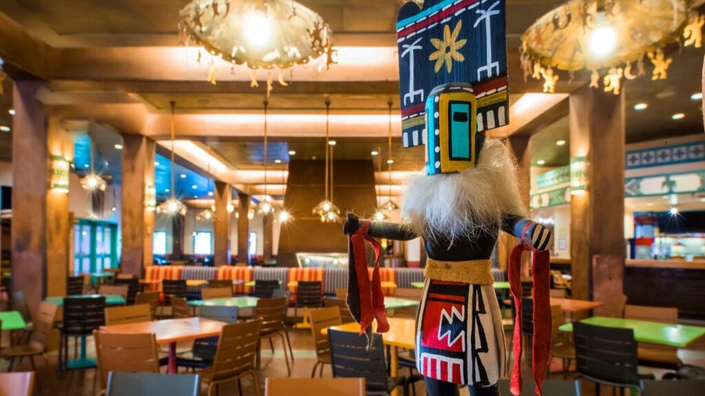 L'intérieur d'un restaurant du Disney Hotel Santa Fe présente une grande figure de style indigène avec une coiffe colorée, entourée de tables et de chaises vides sous des luminaires décoratifs.