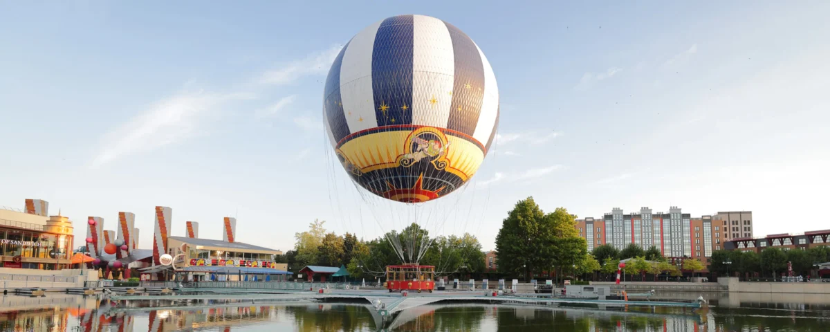 Une montgolfière captive décorée d'un motif de cirque plane au-dessus d'un étang près d'arbres, de bâtiments et d'un petit parc d'attractions à Disney Village, offrant une expérience magique à tous les visiteurs.
